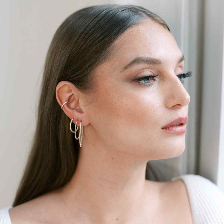 A model wears three diamond hoops with round brilliant diamonds, along with a thin ear cuff with diamonds. The earrings here are sizes 16, 22, and 30mm - stacked on the same ear with the cuff in Yellow Gold.