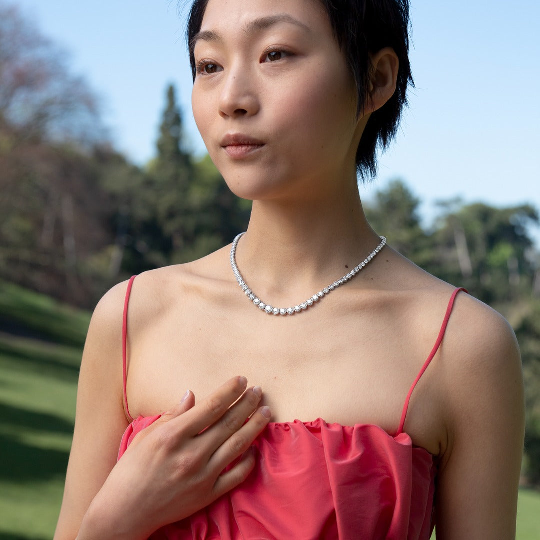 A model wears the Collier Rivière Tennis Necklace as an addition to a springy cocktail look. This 25ct graduated diamond tennis necklace features flawless round brilliants in a four prong solitaire setting. D-color, IF/VVS diamonds crescendo toward a center diamond of 1ct. The length of this striking piece is 16 inches (40cm).