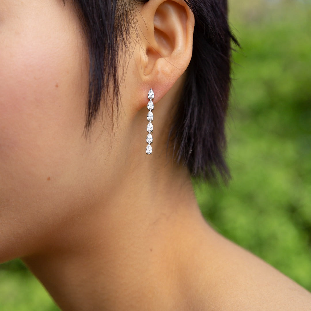 A model wears the striking Île Column Grande Earrings - which shine on their own. Hand-cast from 3.6 grams of recycled 18k gold, these pear diamond earrings feature six pristine tear drops floating in an architectural column. The pair of earrings is 2 total carat weight. The shape and size is fully customizable.