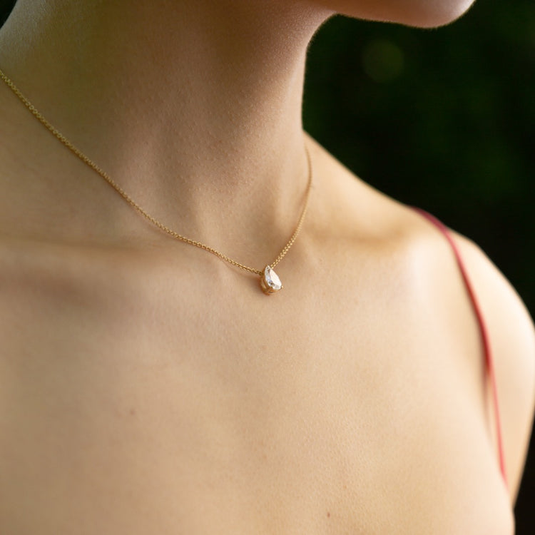 A model wears the Île Pear Necklace with a Yellow Gold setting and chain. A solitaire pear diamond is centered between the collarbone on an 18k gold chain. The length of this necklace is 16" (40cm). Please get in touch if you would like a different chain length. Shape, color, and size of this pear diamond necklace are fully customizable as well.