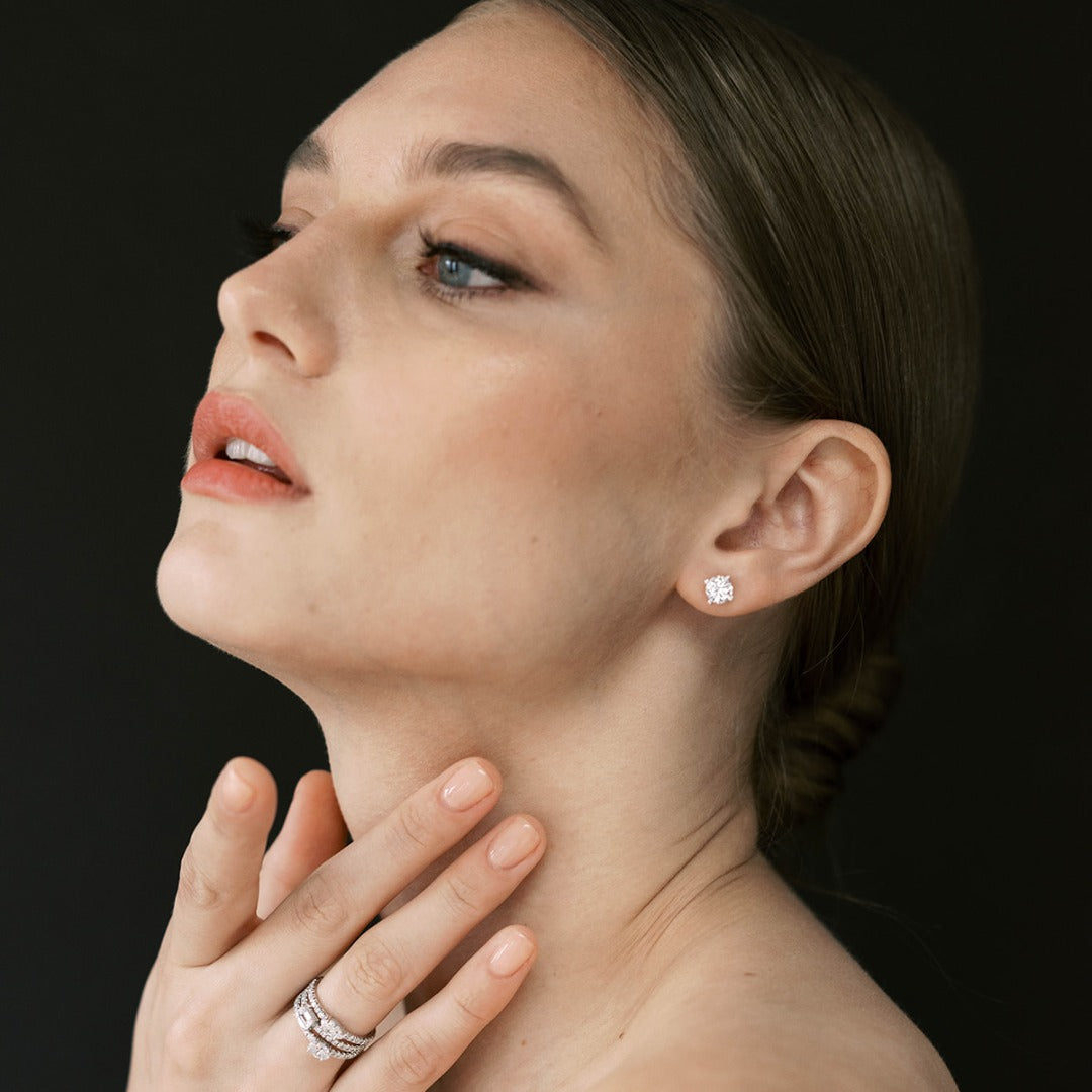 A model wears the Île Studs in a wedding gown. Polished 18k gold is illuminated by a solitaire D color, IF/VVS diamond, the unparalleled quality of which is on full display in a 4-prong setting. Size and shape customizable. Wearing the earrings with the Trois Pierres Rings.