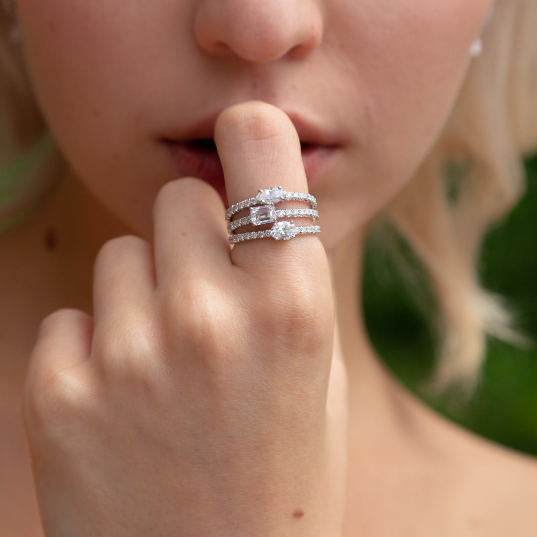 A model wears the Trois Pierres set - three rings of varying diamond shapes paired together. Diamond center shapes from top ring to bottom: marquise, emerald, and oval, with pavé diamonds around the entire band. The center diamonds is fully customizable to any shape and any pairing.